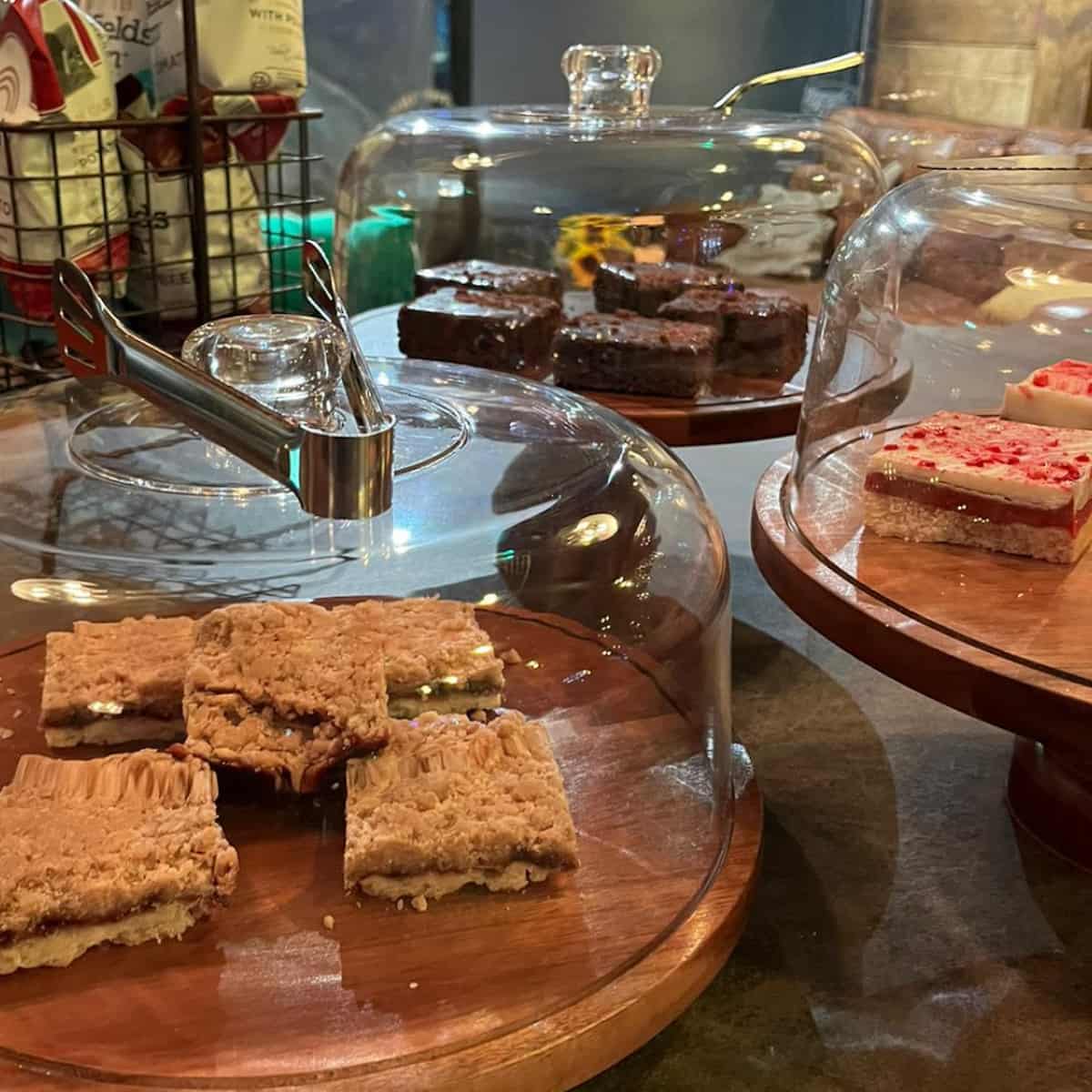 Slices of cake, flapjack and shortbread on wooden platters, covered with glass domes.