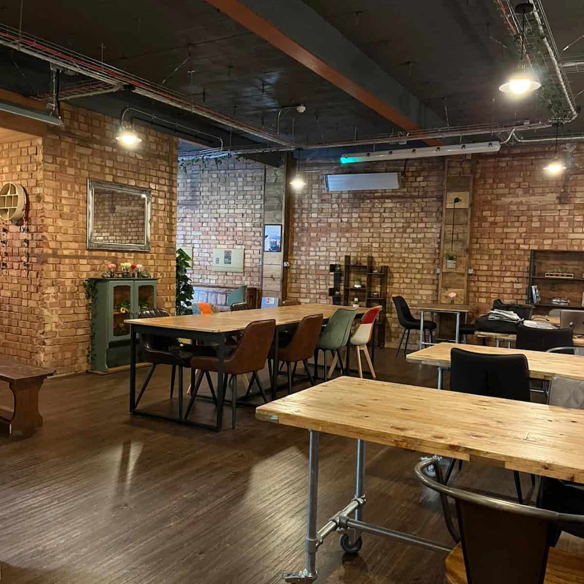 Several long wood-topped tables surrounded by chairs, on a brown floor and with a brick wall, shelves and a mirror in the background, in the first floor at Understated coffee shop. 
