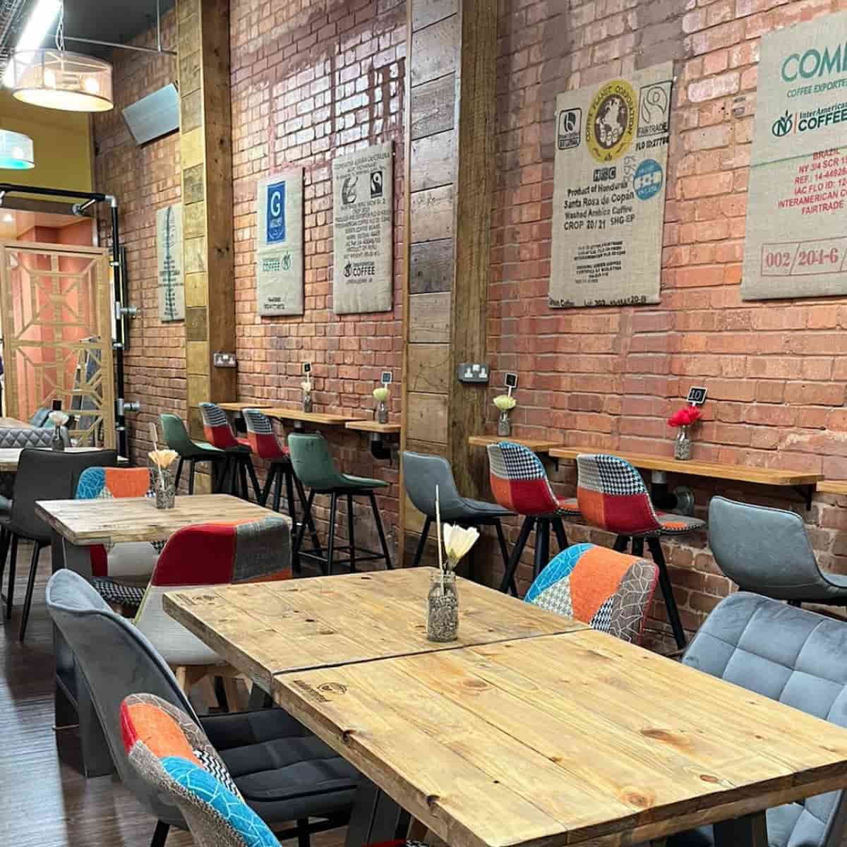 Wood-topped cafe tables in Understated coffee shop, surrounded by orange, turquoise and black and white pattern chairs. In the background, a plain brick wall and pictures made from coffee sacks
